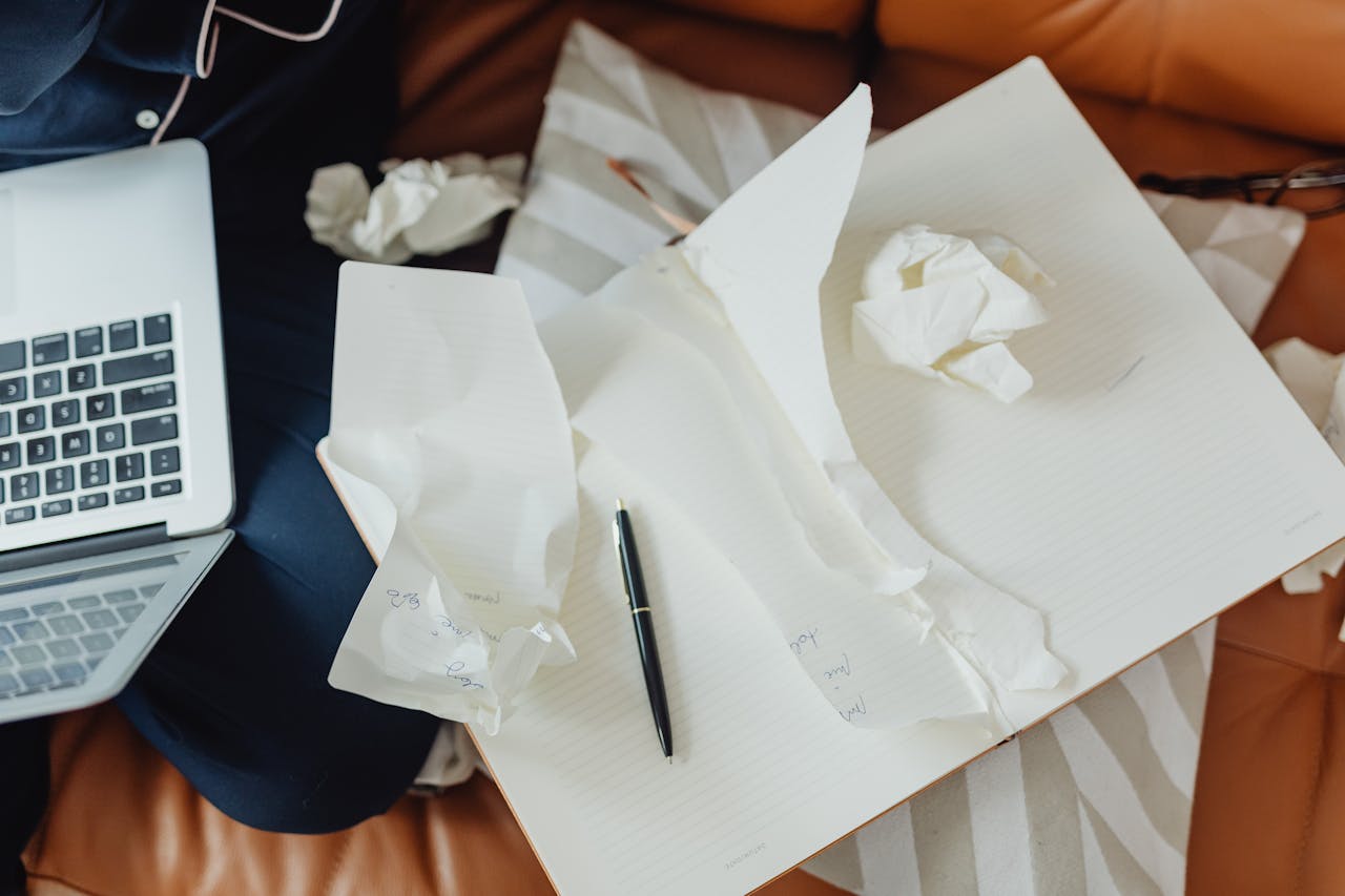 A creative workspace featuring a laptop, crumpled papers, and an open notebook with a pen, showcasing brainstorming or writing activities.